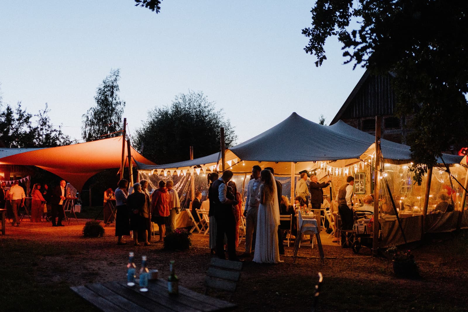Brautpaar und Gäste vor den mit Lichterketten beleuchteten Stretchzelten in der Abenddämmerung bei der Hochzeit von Lea und Malte auf dem Hof Eggers in der Ohe